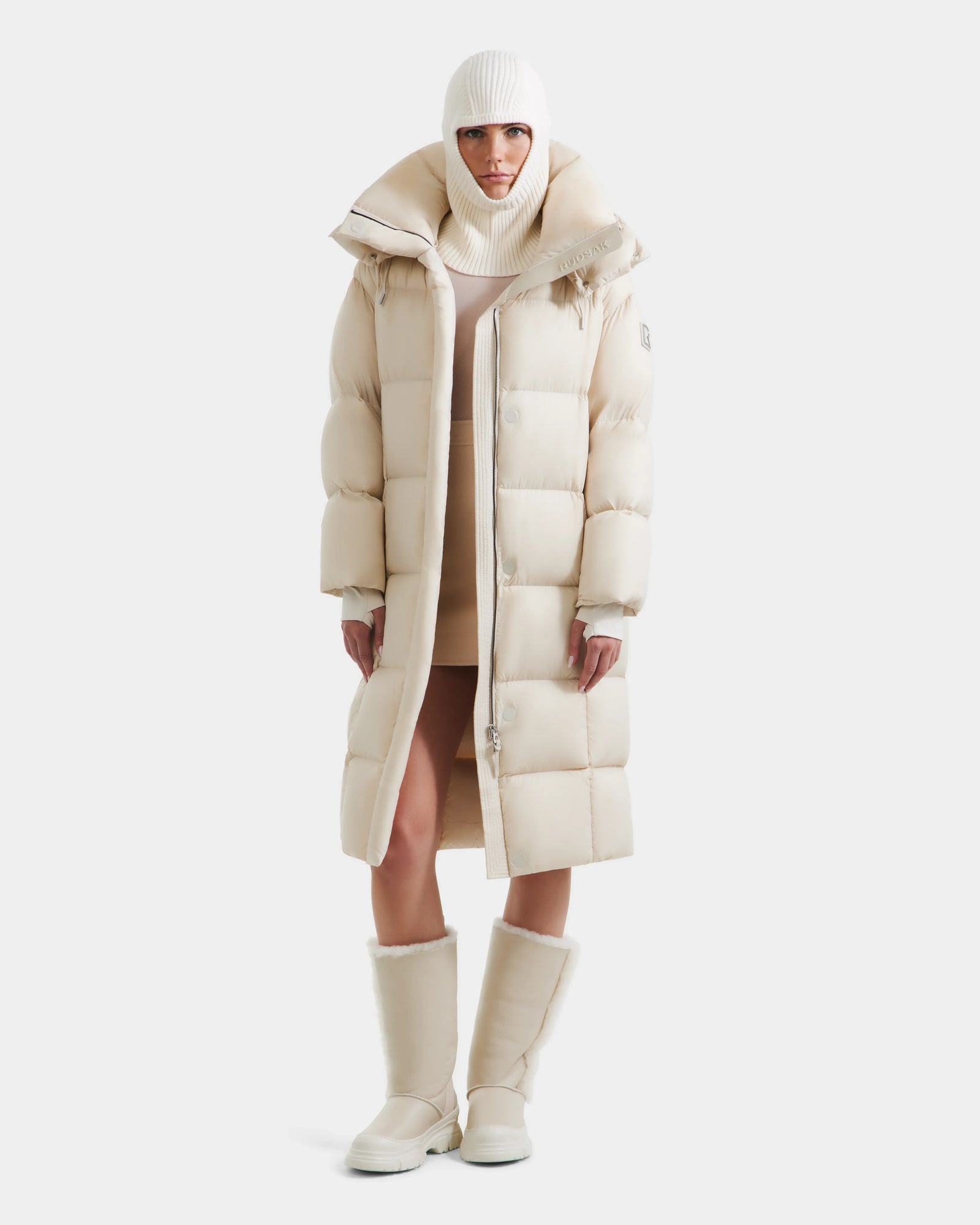 A woman faces forward wearing the Rudsak SASHA - 8125686 Vanilla, a women’s long down puffer coat with removable hood, styled with a matching knit hat and boots—all in soft off-white—set against a clean white background.