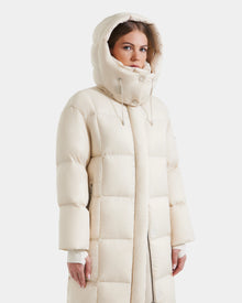 A woman models the Rudsak SASHA - 8125686 Vanilla women's long down puffer coat in cream with a high collar and hood, offering stylish warmth for winter, shown on a plain white background.