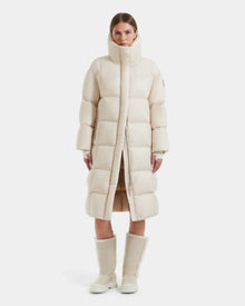 A woman faces forward wearing the Rudsak SASHA - 8125686 Vanilla long down puffer coat with a high collar and removable hood, paired with winter boots. A plain white background emphasizes the sleek cream style.