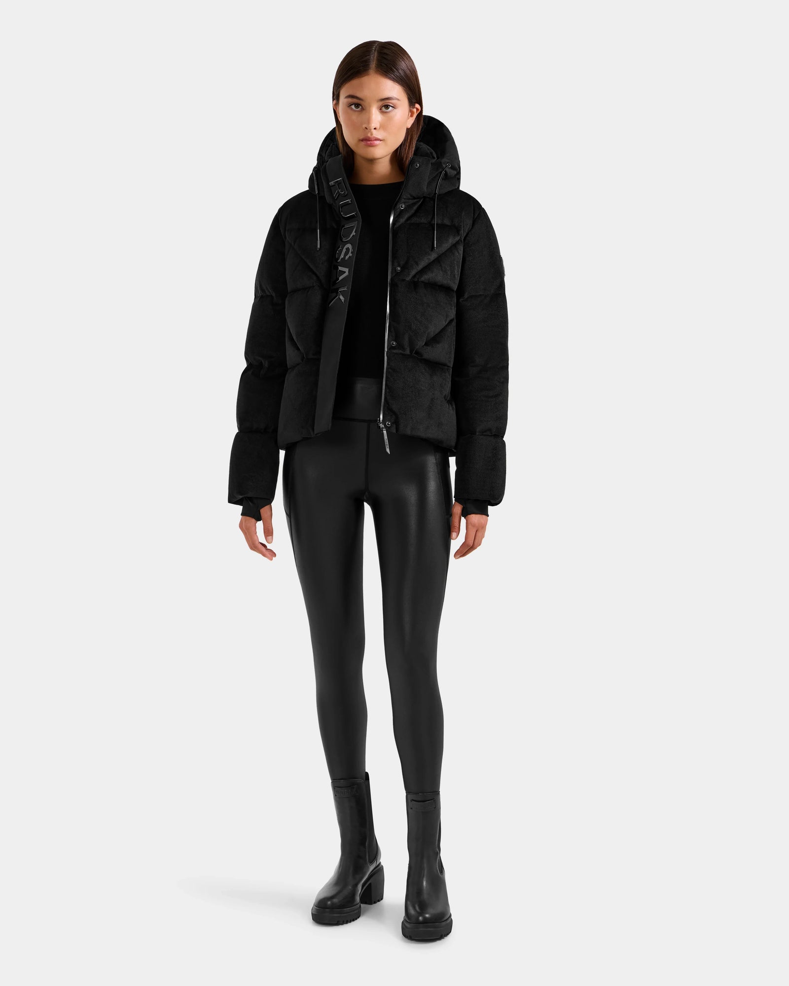 A woman faces forward, wearing the Rudsak STEFANIE V - L125584 Black down puffer winter jacket with a hood, paired with black fitted leggings and ankle boots. Her hands rest at her sides against a plain light gray background.
