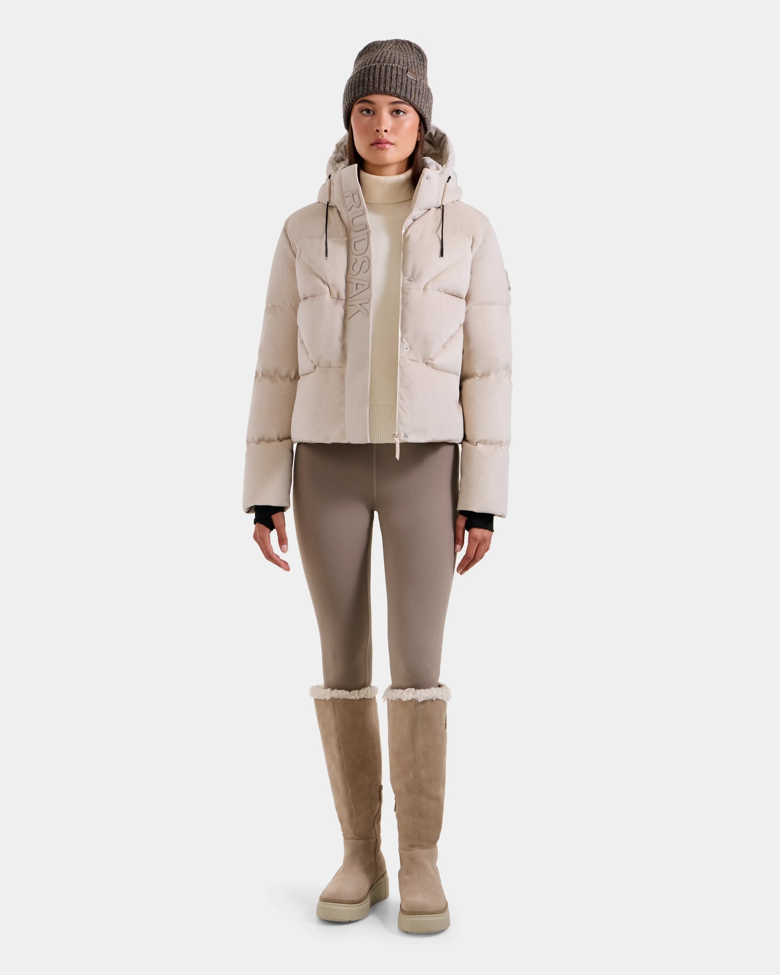 A woman stands facing forward in the STEFANIE V - L125584 Vanilla down puffer jacket by Rudsak, paired with tan leggings, tall tan boots with fleece trim, a cream turtleneck, and a gray knit beanie against a plain white background.