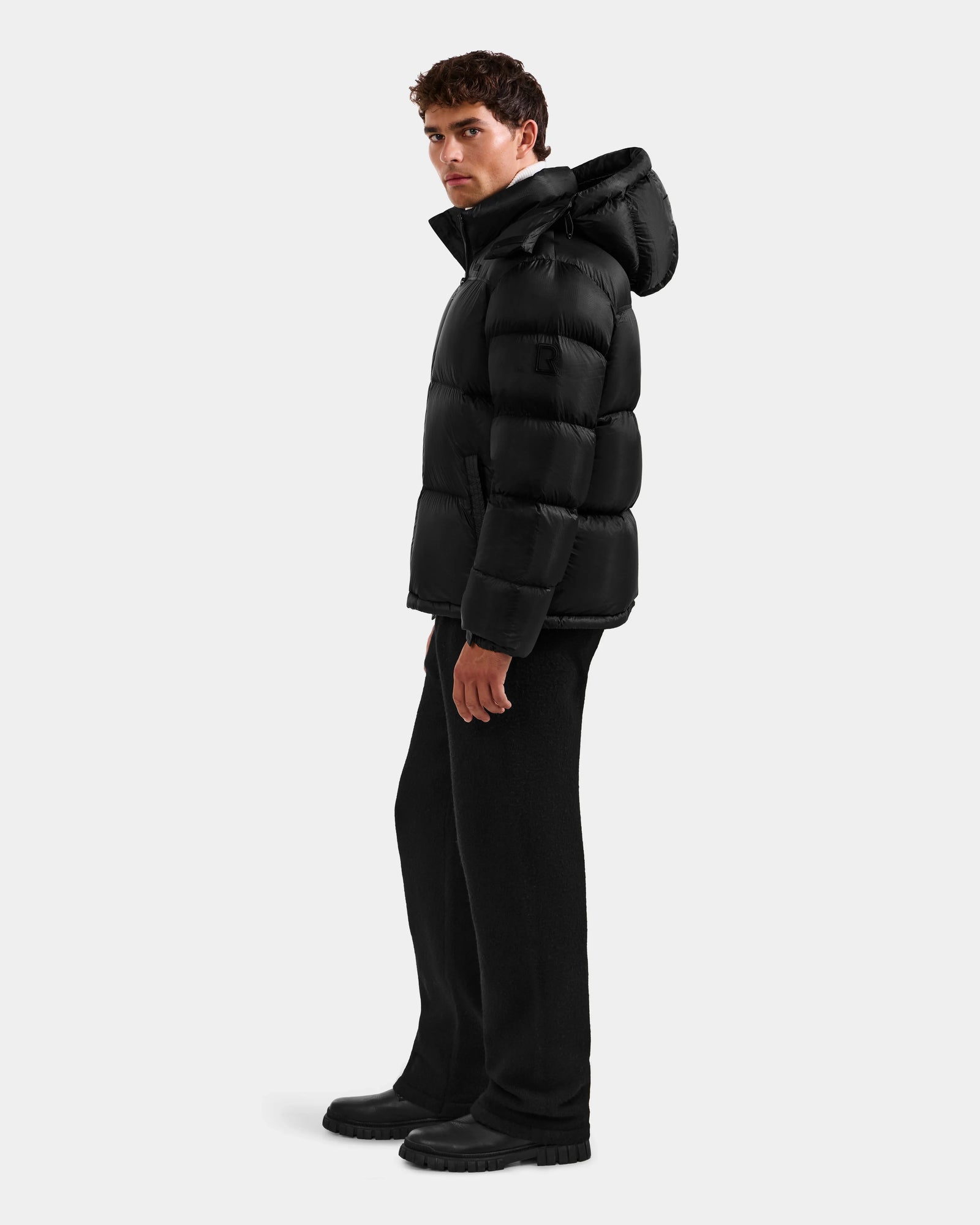 A man wearing the Rudsak STEFANO - 6125574 Black men’s down puffer, paired with black pants and boots, stands against a plain light gray background, facing slightly to the side.