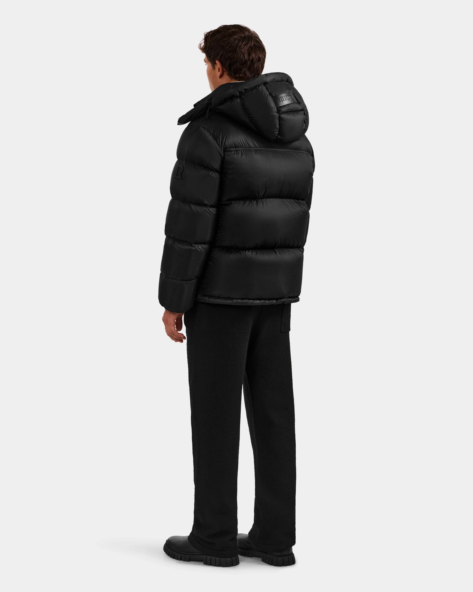 A person faces away, wearing the Rudsak STEFANO - 6125574 Black men’s down puffer jacket with a hood, black pants, and black shoes, set against a plain light gray background.