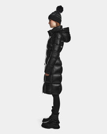 A person stands in profile against a plain white background, wearing the Rudsak VENICE S - L124514 Black women's winter coat with a hood, paired with black leggings, chunky black boots, and a black knit beanie with pom-pom.