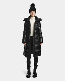 A woman wearing the Rudsak VENICE S - L124514 Black winter coat, a black pom-pom knit hat, white turtleneck, black leggings, and boots stands facing forward against a plain white background. The quilted puffer features RDS-certified down.