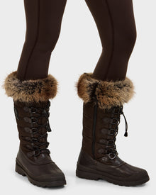 A person stands on a light background wearing dark brown leggings and Rudsak WINDY - L224507 Espresso women’s winter boots, featuring faux fur trim and lace-up fronts—ideal for cold-weather comfort.