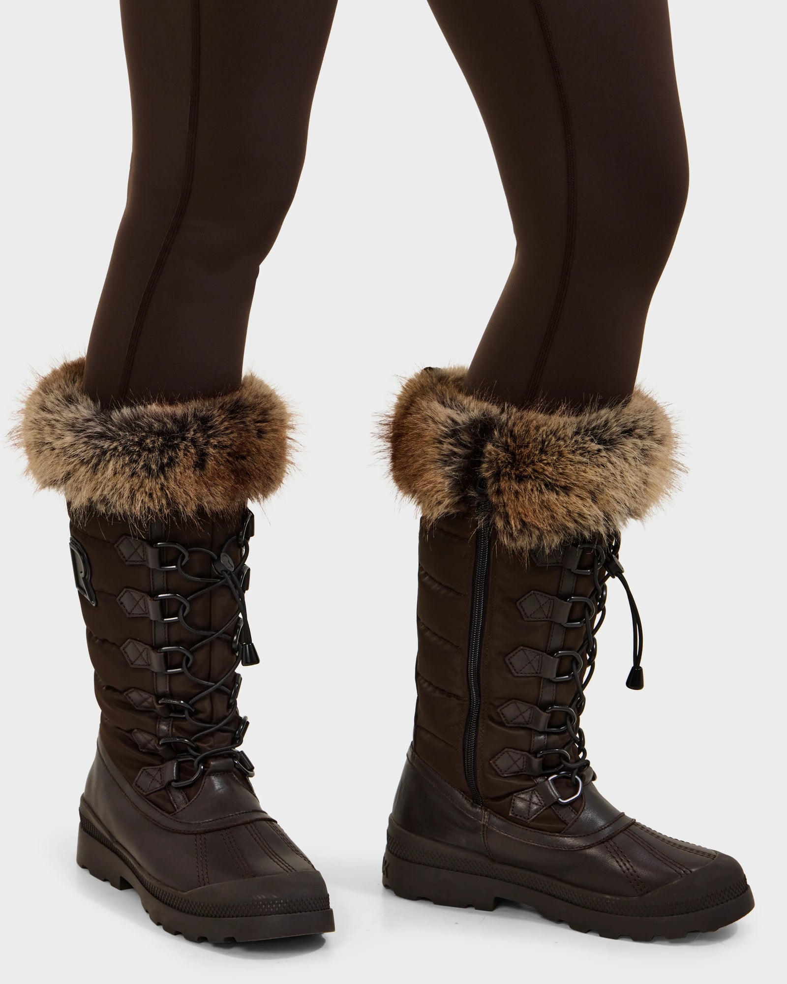 A person stands on a light background wearing dark brown leggings and Rudsak WINDY - L224507 Espresso women’s winter boots, featuring faux fur trim and lace-up fronts—ideal for cold-weather comfort.