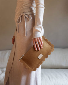A person in a light beige dress with a waist tie holds a Graf Lantz versatile clutch featuring scalloped edges. Rings adorn their hand, while the scene includes a light-colored sofa against a neutral wall.