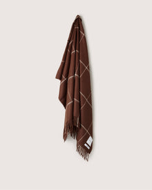 A brown fringed throw blanket by Graf Lantz, crafted from naturally-dyed baby alpaca wool with a white windowpane pattern, hangs on a light-colored wall. Made in Peru.