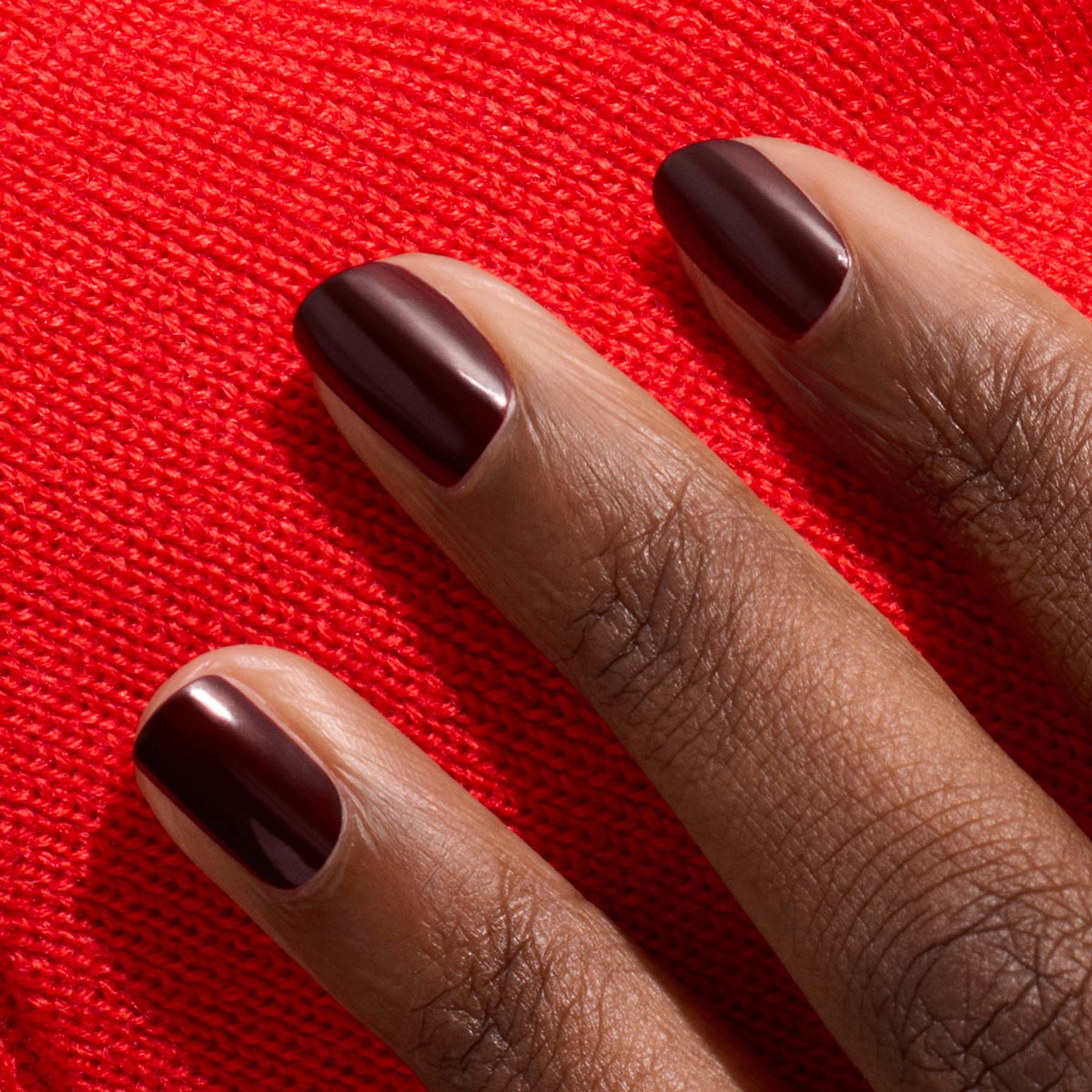 Three neatly manicured short nails in Manucurist's Hollyhock, a deep red polish, rest against vibrant red fabric, complementing a rich skin tone.