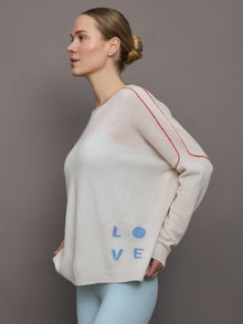 Model wears a white sweater with red seams along the biceps and blue block lettering on front bottom right that says LOVE. Back of sweater says in blue lettering 
