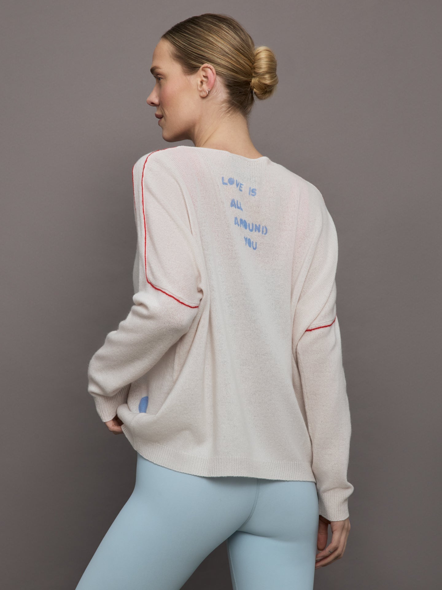 Model wears a white sweater with red seams along the biceps and blue block lettering on front bottom right that says LOVE. Back of sweater says in blue lettering "Love is all around you."