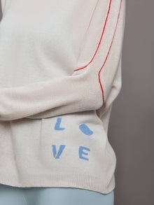 Model wears a white sweater with red seams along the biceps and blue block lettering on front bottom right that says LOVE. Back of sweater says in blue lettering 