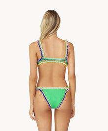 A blonde woman wearing a green bikini facing towards a white wall. 