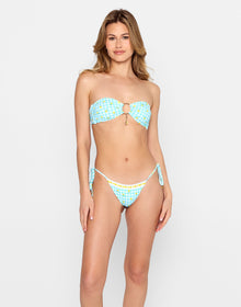 Model posing with her back to the camera in the 'Limoncello Spritz Thong Bottom - Limoncello' bikini, featuring a cheeky fit in a blue gingham print with lemon details. She stands against a light blue brick wall, holding a drink and soaking up the sun.