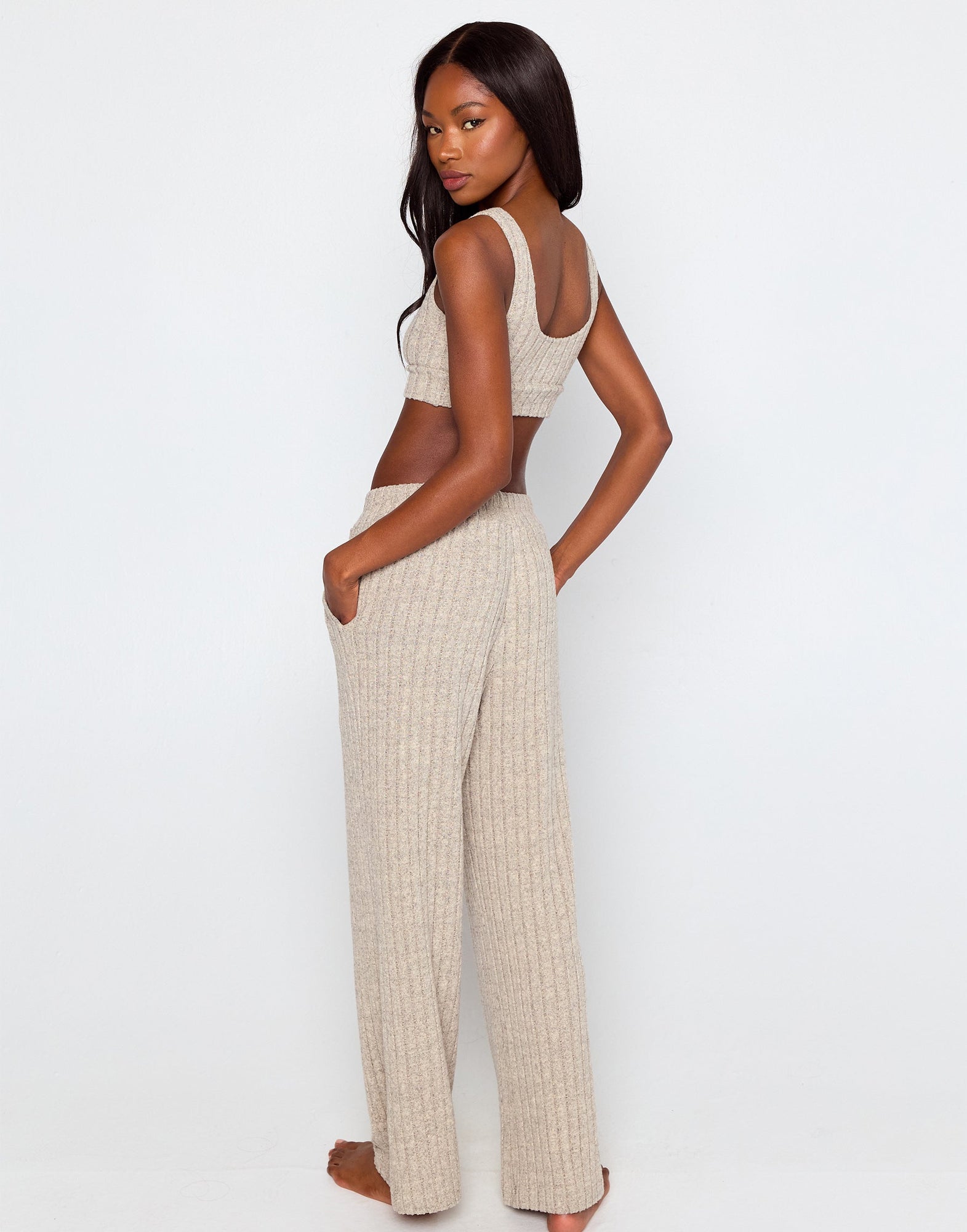 Woman wearing a beige ribbed knit loungewear set, featuring a cropped tank top with a scoop neckline and wide-leg drawstring pants with side pockets, standing barefoot against a plain white background.