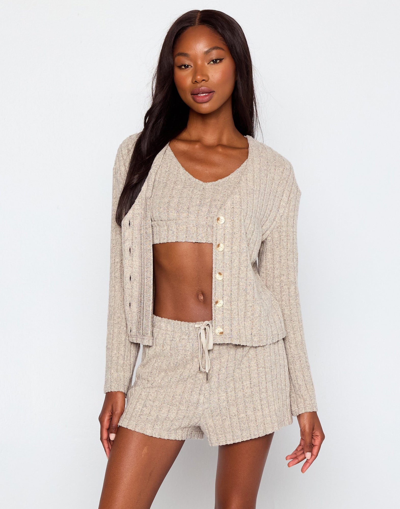 Woman wearing a beige ribbed knit loungewear set including a cropped tank top, drawstring shorts, and a matching button-up cardigan with long sleeves, posing against a plain white background.