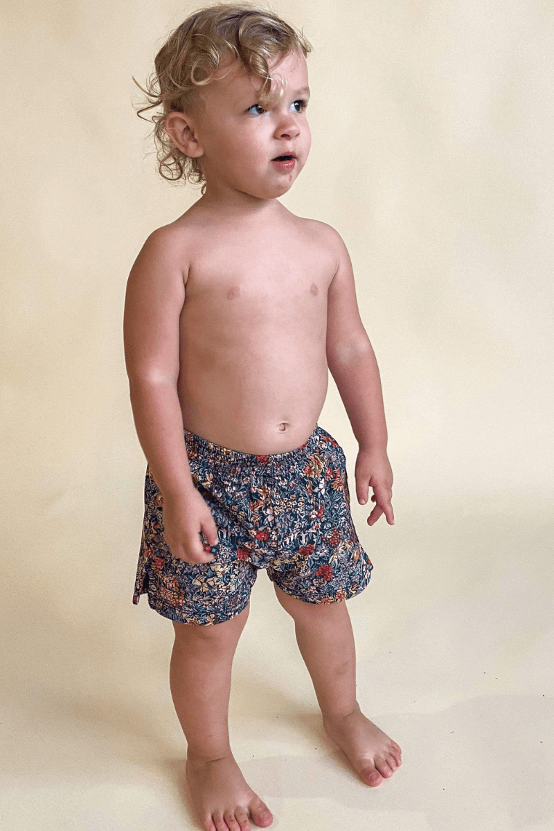 Toddler wearing navy swim trunks with multi-colored floral print, shown from side profile.