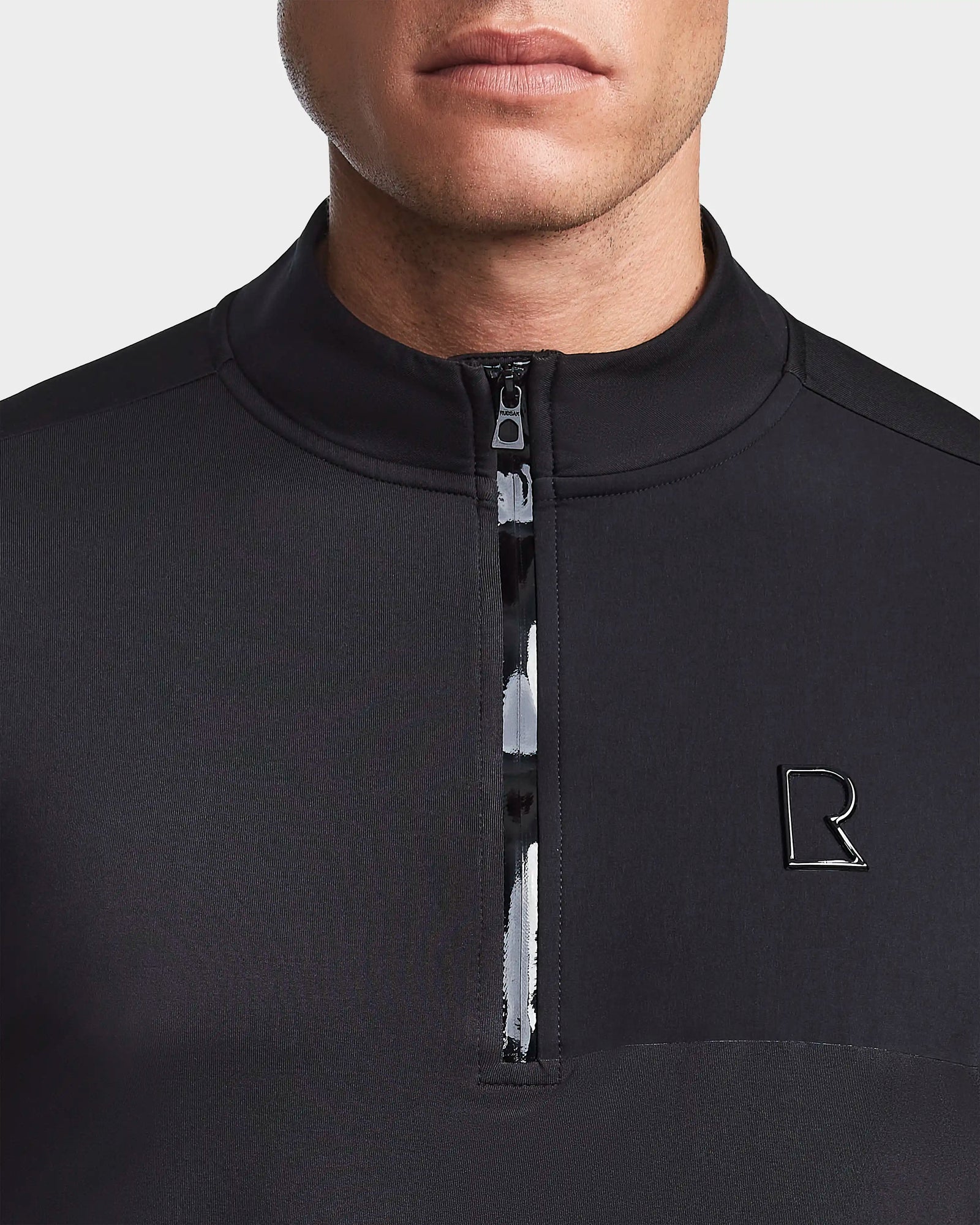 A close-up shows a man in the Rudsak WARREN - 6523502 Black zip-front mock neck, featuring a small metallic R logo on the chest. Only his lower face and upper torso are visible in this sleek mens fitted stretch top.
