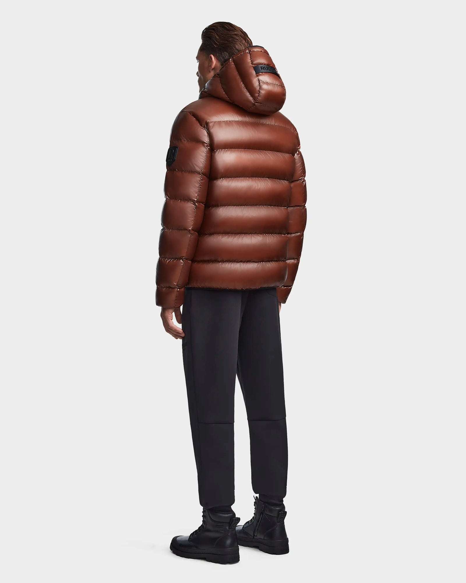 A person faces away, wearing the Rudsak DEMIAN S - M123508 Cohiba shiny brown hooded down puffer jacket, which is water- and wind-resistant, paired with black pants and boots against a plain light background.