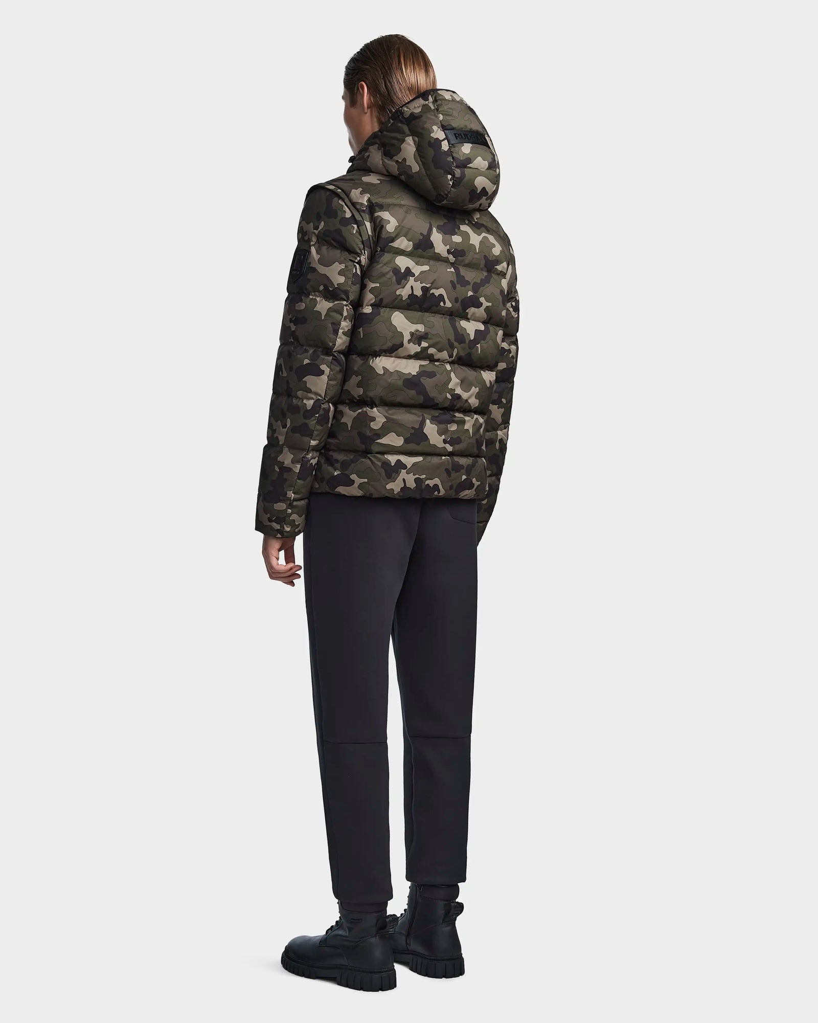 A person faces away, wearing the Rudsak DEMIAN M-R - M123504 Camouflage mens down puffer with a hood, paired with black pants and boots, set against a plain light background.