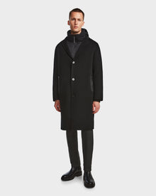 A man faces forward against a white background, wearing Rudsaks DRAL - 6124503 Black 2-in-1 jacket with a removable hooded down puffer, dark pants, and black boots.