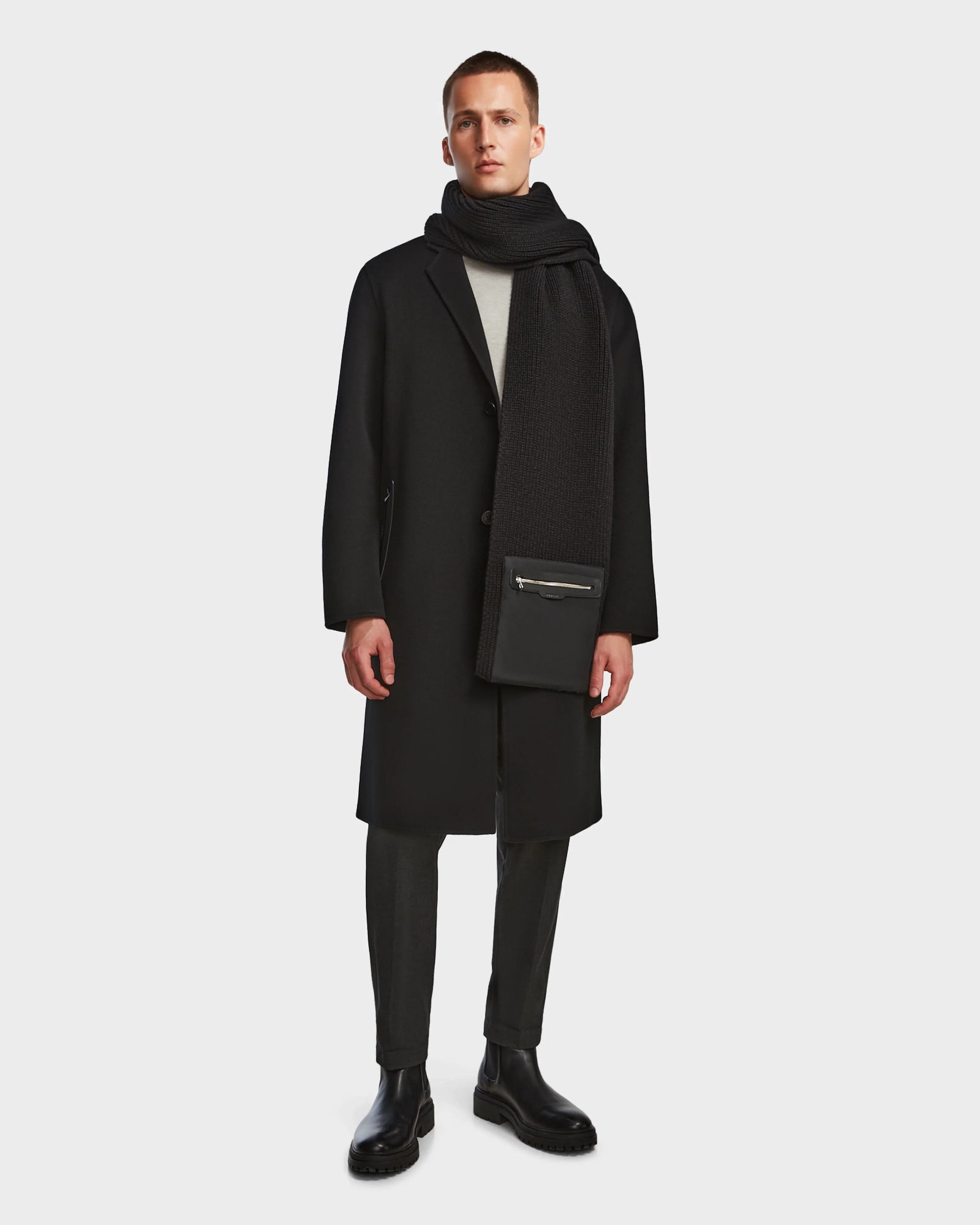 A man stands against a plain background wearing the Rudsak DRAL - 6124503 Black wool jacket styled as a long coat, paired with a black scarf, gray trousers, black boots, and holding a black zippered pouch.