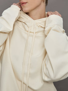 Model wears an oversized cream colored hoodie with extra long drawstrings. 