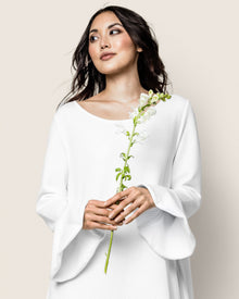 A woman in a Petite Plume Womens Pima Ophelia Nightgown in White Jacquard holds a single white flower, gazing slightly to the side against a light beige backdrop, her dark hair softly draping over her shoulders like Peruvian Pima cotton.