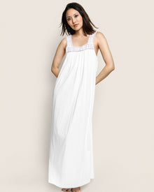A woman poses against a plain background wearing the Womens Pima Camille Nightgown in White by Petite Plume, featuring a decorative lace neckline. Her hands rest behind her back, and her hair is down, softly blowing to one side.