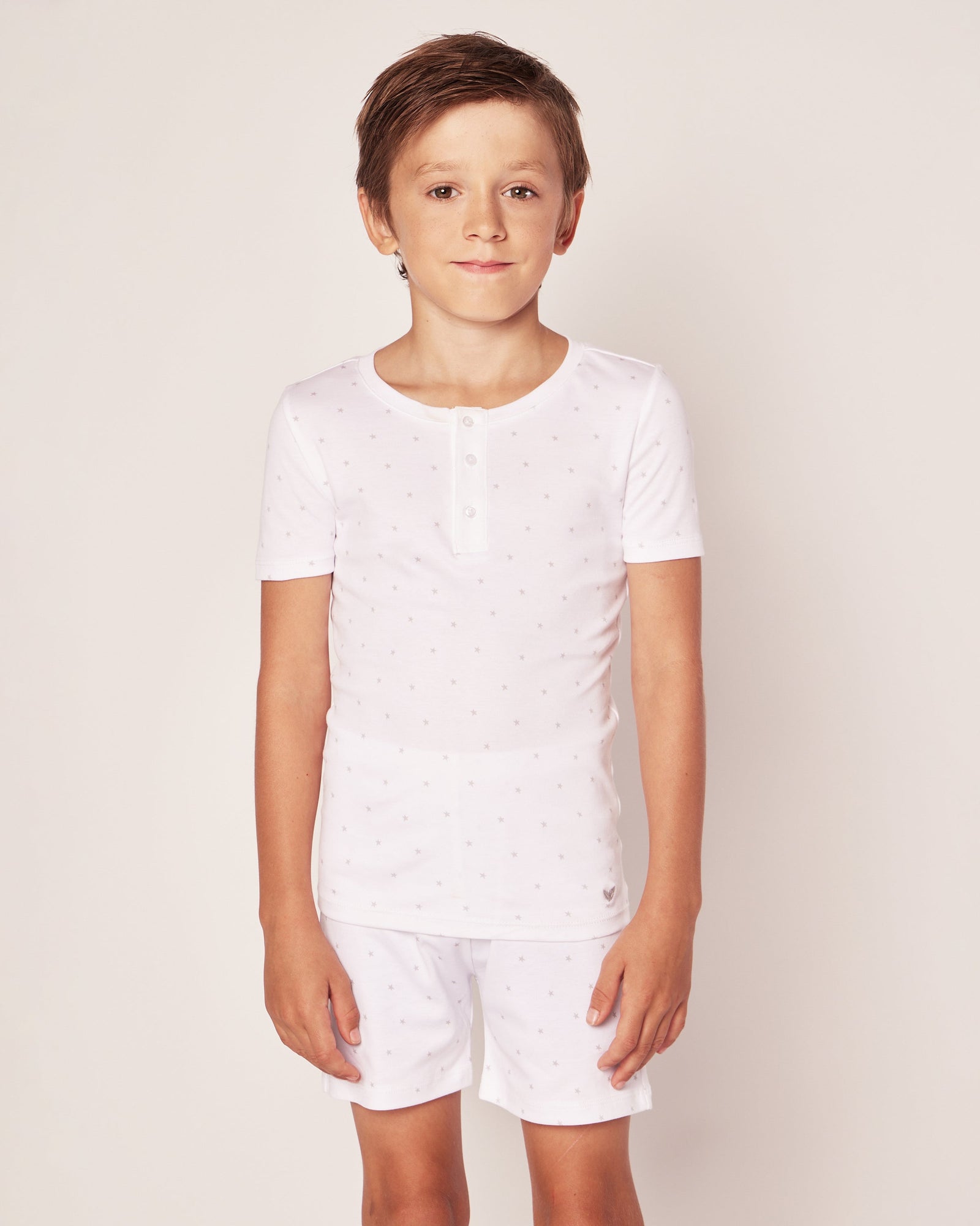 A young boy in Petite Plumes Kids Pima Snug Fit Pajama Short Set in Grey Stars, featuring a white short-sleeved shirt with subtle patterns and matching shorts, stands against a plain background, smiling at the camera.
