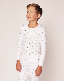A child in Petite Plumes Kids Pima Snug Fit Pajama Set in Evergreen Trees, made of soft Pima cotton, stands against a plain background looking to the side. The long sleeve top and pant set exudes a light and neutral tone.