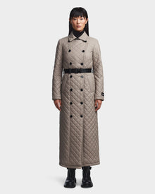 A woman faces forward in the Rudsak DAISY - 8123580 Cement women’s quilted maxi trench coat with sustainable Sorona insulation, double-breasted buttons, black belt, diamond pattern to the ankles, and black boots against a white background.