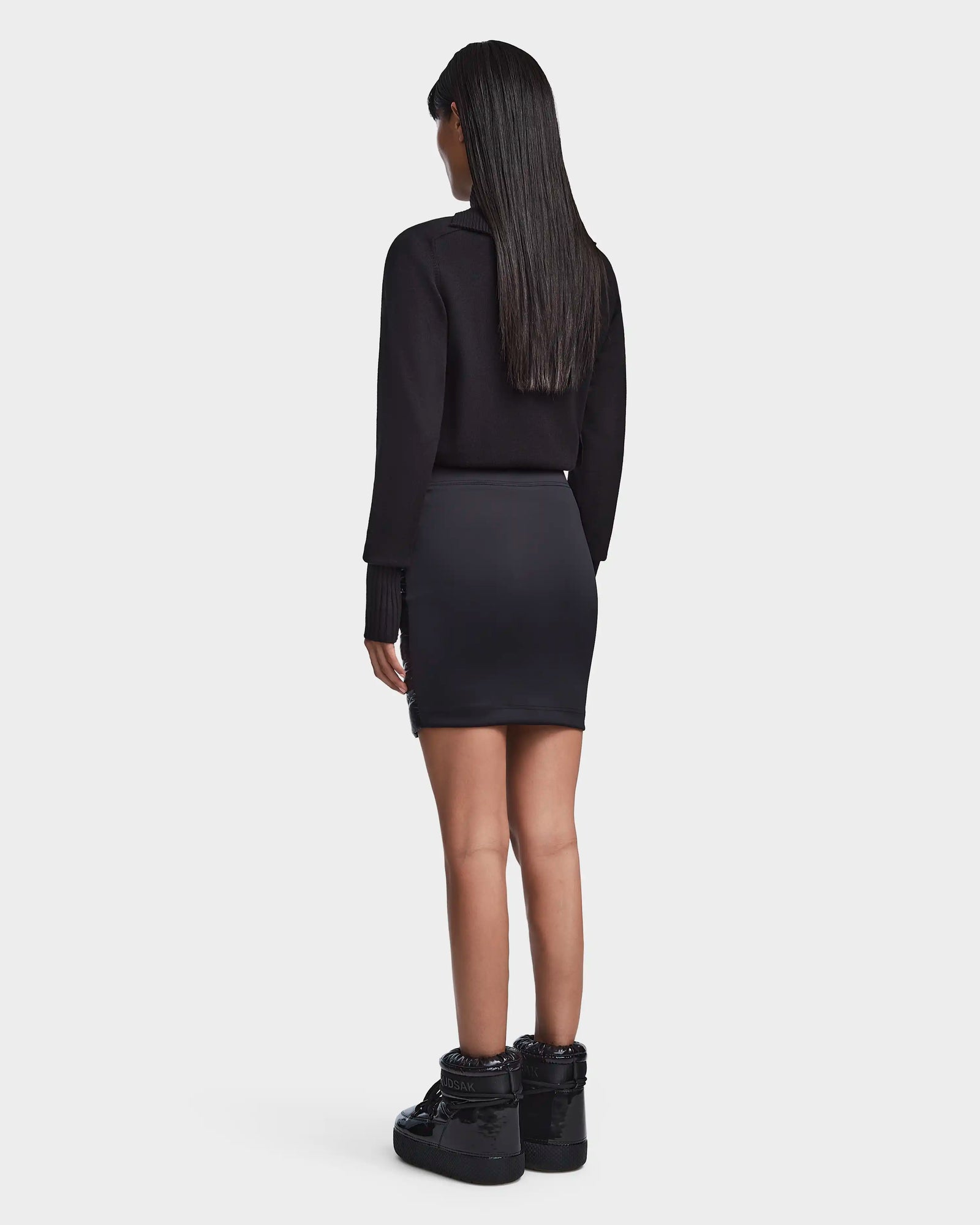 A woman with straight, long dark hair stands facing away in a black Rudsak MARZI - 8523528 chevron quilted winter skirt, paired with a black long-sleeve top and black lace-up boots against a plain white background.