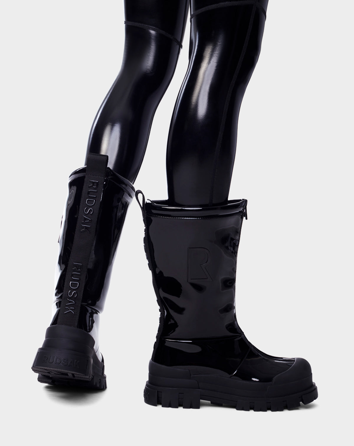 A person wears Rudsaks KRISTAL HI - 8222510 BLACK womens boots, featuring high-gloss finish, chunky grip soles, and RUDSAK branding at the back, standing against a plain light background.