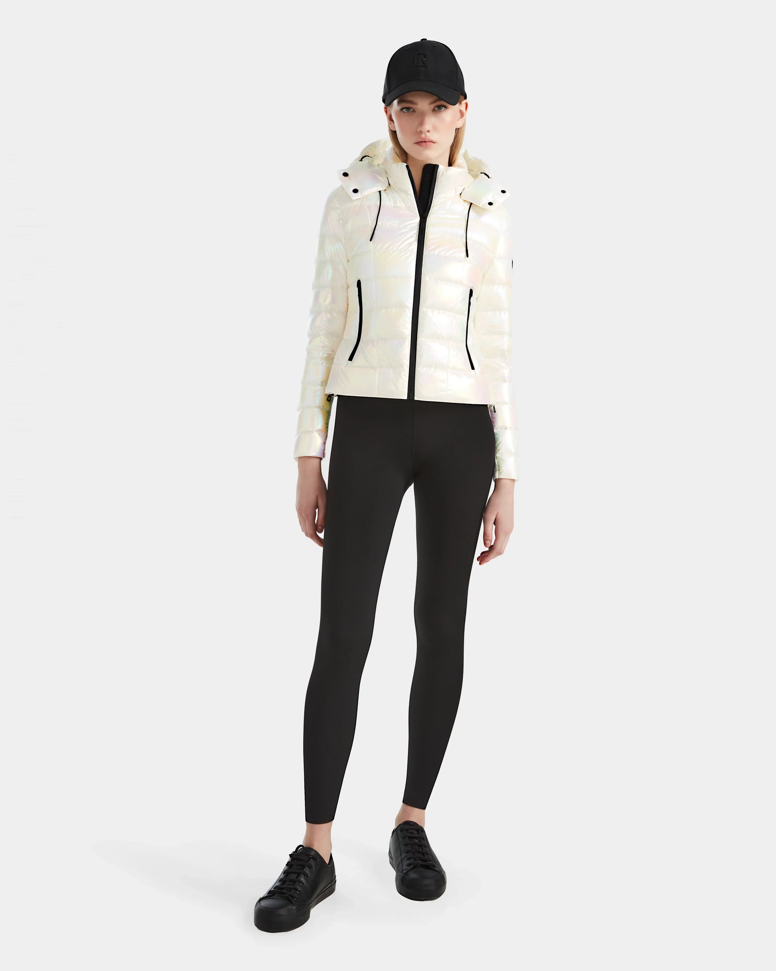 A woman stands against a plain background wearing the Rudsak MELISSA - 8124031 Iridescent Eggshell lightweight down puffer jacket, black leggings, sneakers, and a black baseball cap. She has a neutral expression with arms relaxed at her sides.