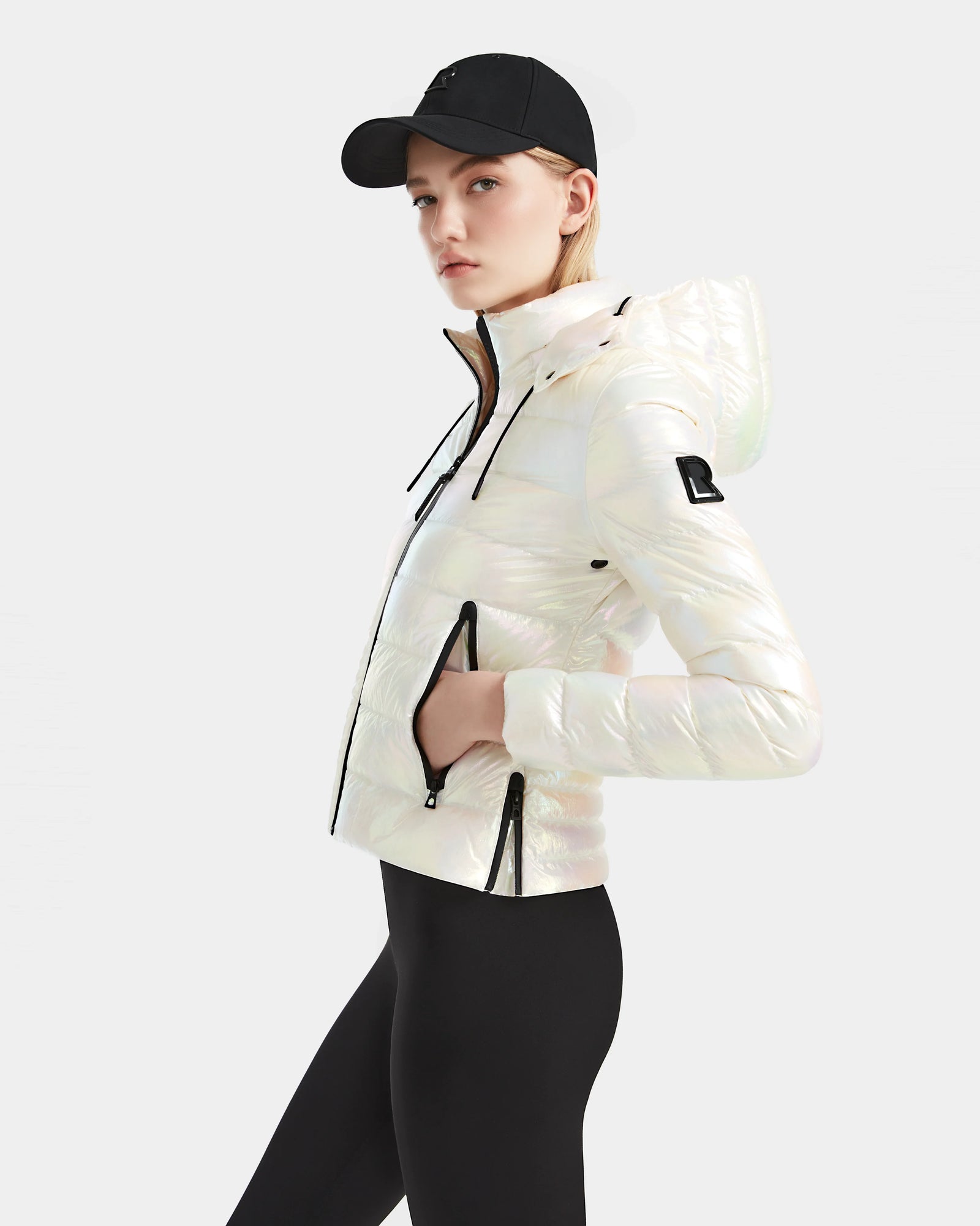 A woman models the Rudsak MELISSA - 8124031 Iridescent Eggshell weatherproof down jacket with a hood, styled with black leggings and a black baseball cap against a plain light background, facing slightly to the side.