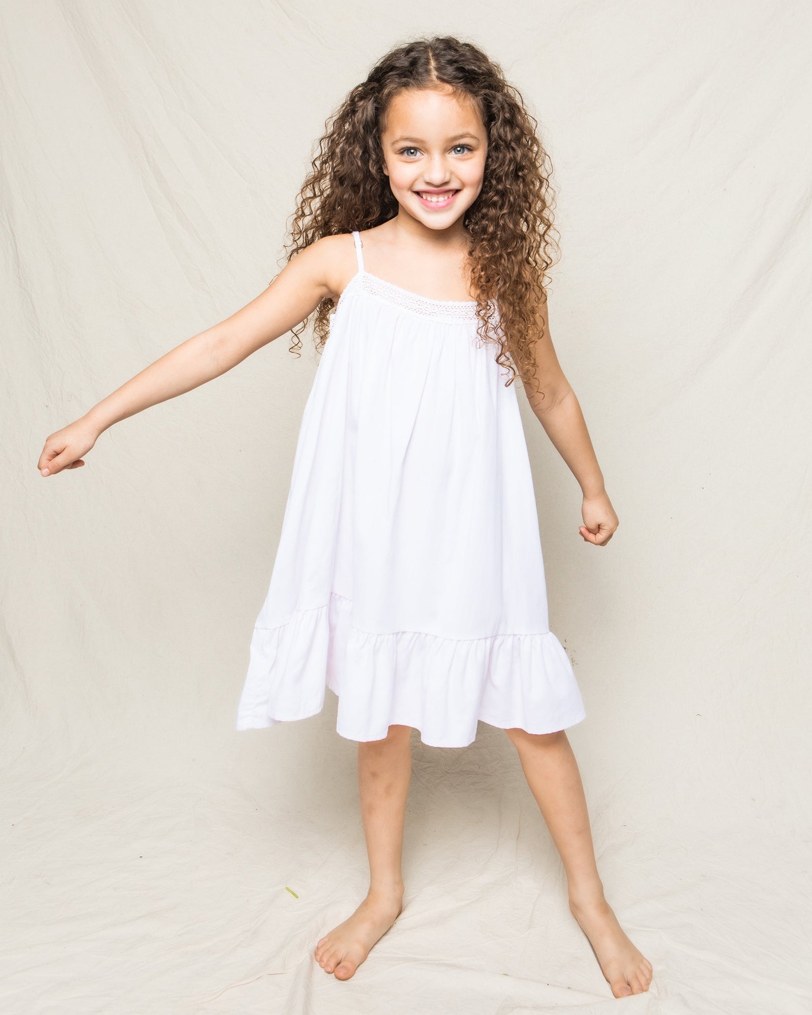 A cheerful young girl with curly hair wears Petite Plumes Girls Twill Lily Nightgown in White, its yarn-dyed cotton adding charm as she smiles barefoot against a light backdrop, her right arm slightly raised.