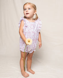 A young child with blonde pigtails stands barefoot against a plain background, wearing Petite Plumes enchanting Babys Twill Ruffled Romper in Dorset Floral, reminiscent of the Victorian era. They hold a yellow daisy and gaze upwards.