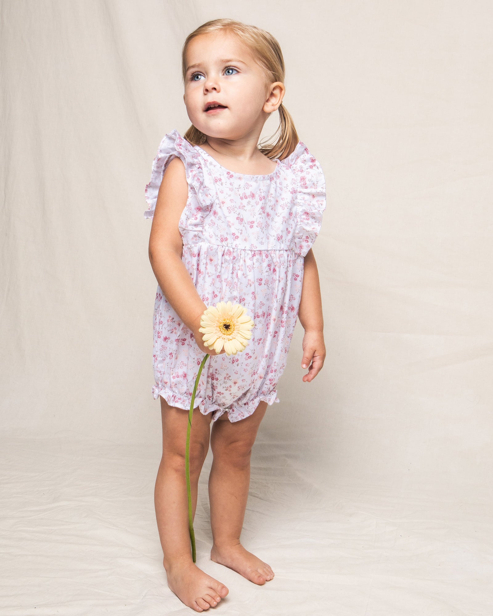 A young child with blonde pigtails stands barefoot against a plain background, wearing Petite Plumes enchanting Babys Twill Ruffled Romper in Dorset Floral, reminiscent of the Victorian era. They hold a yellow daisy and gaze upwards.