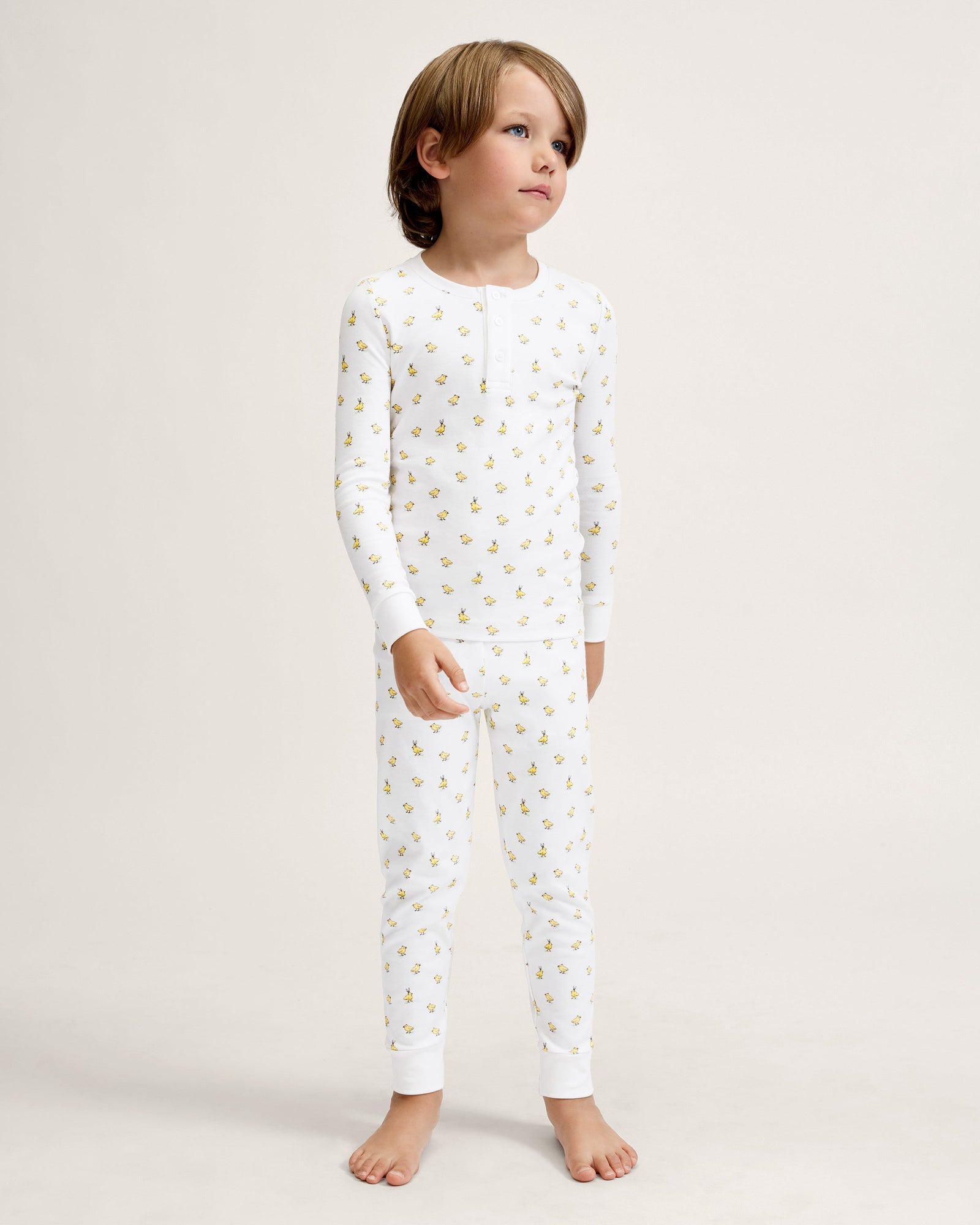 A young child with light brown hair stands barefoot on a plain background, wearing the Kid's Pima Snug Fit Pajama Set in Tres Chick and looking slightly to the side.