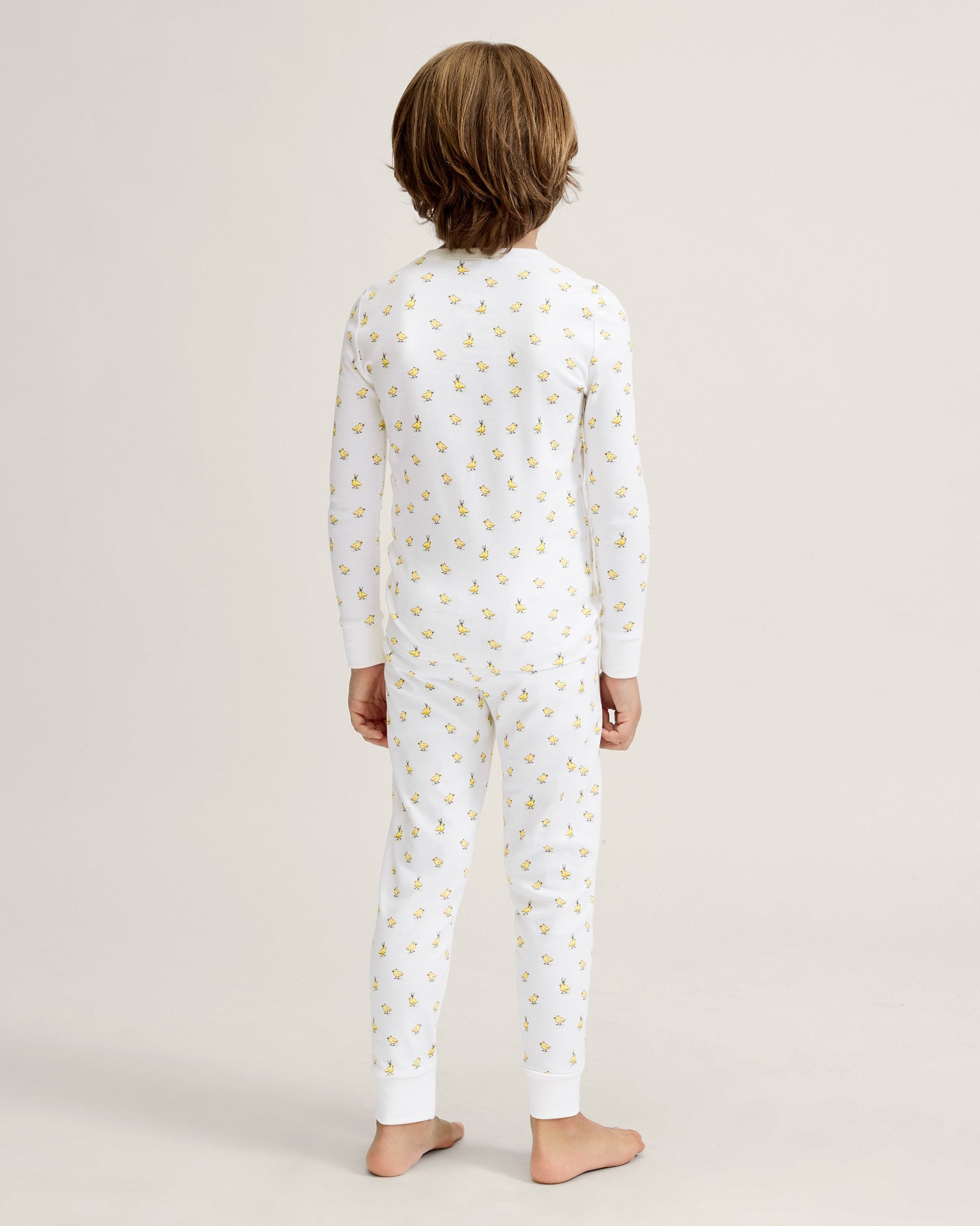 A young child with light brown hair stands barefoot on a plain background, wearing the Kid's Pima Snug Fit Pajama Set in Tres Chick and looking slightly to the side.