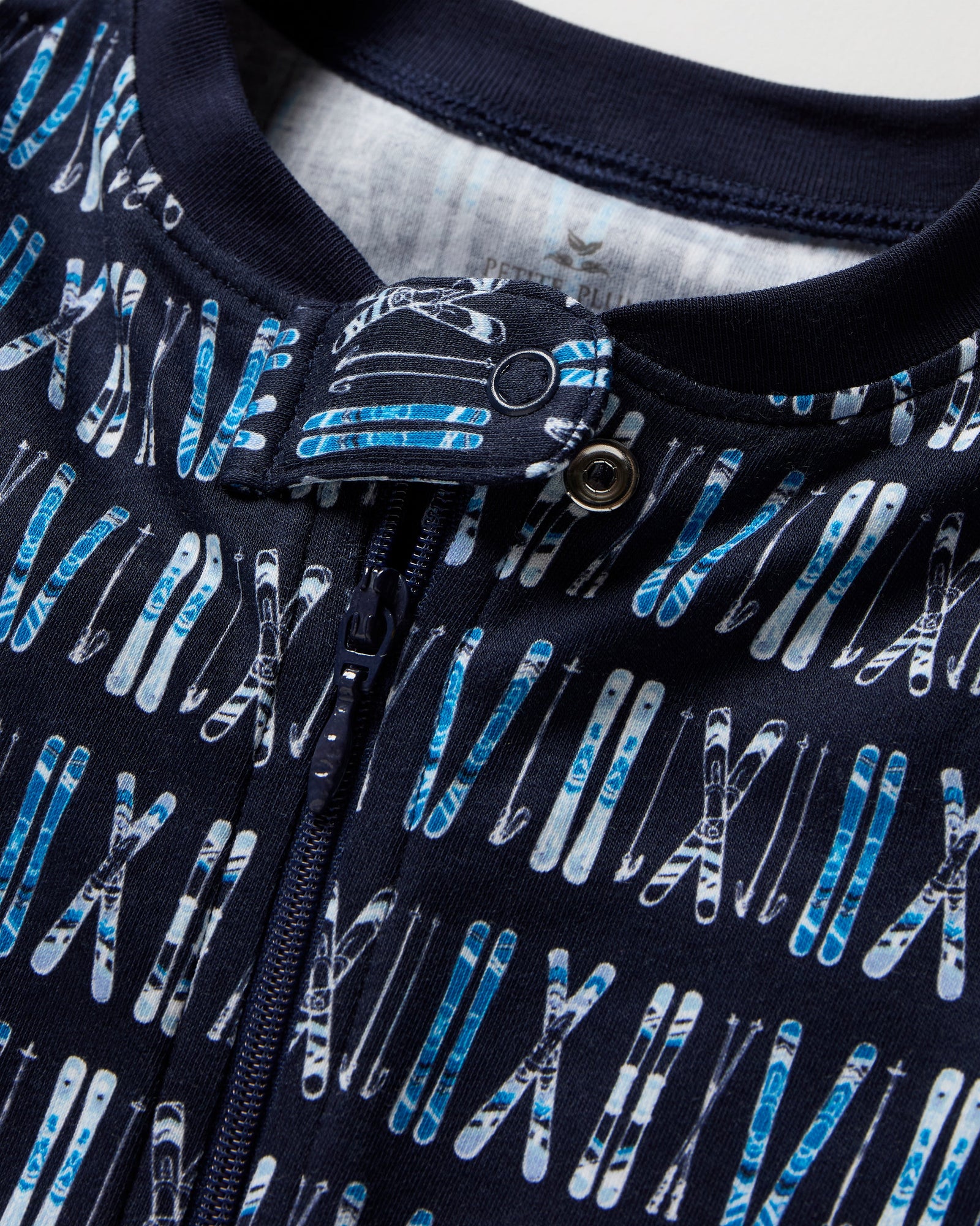The Baby's Pima Romper in Ski Trails is a navy blue zip-up footed onesie with an all-over pattern of white and blue skis and ski poles arranged vertically.