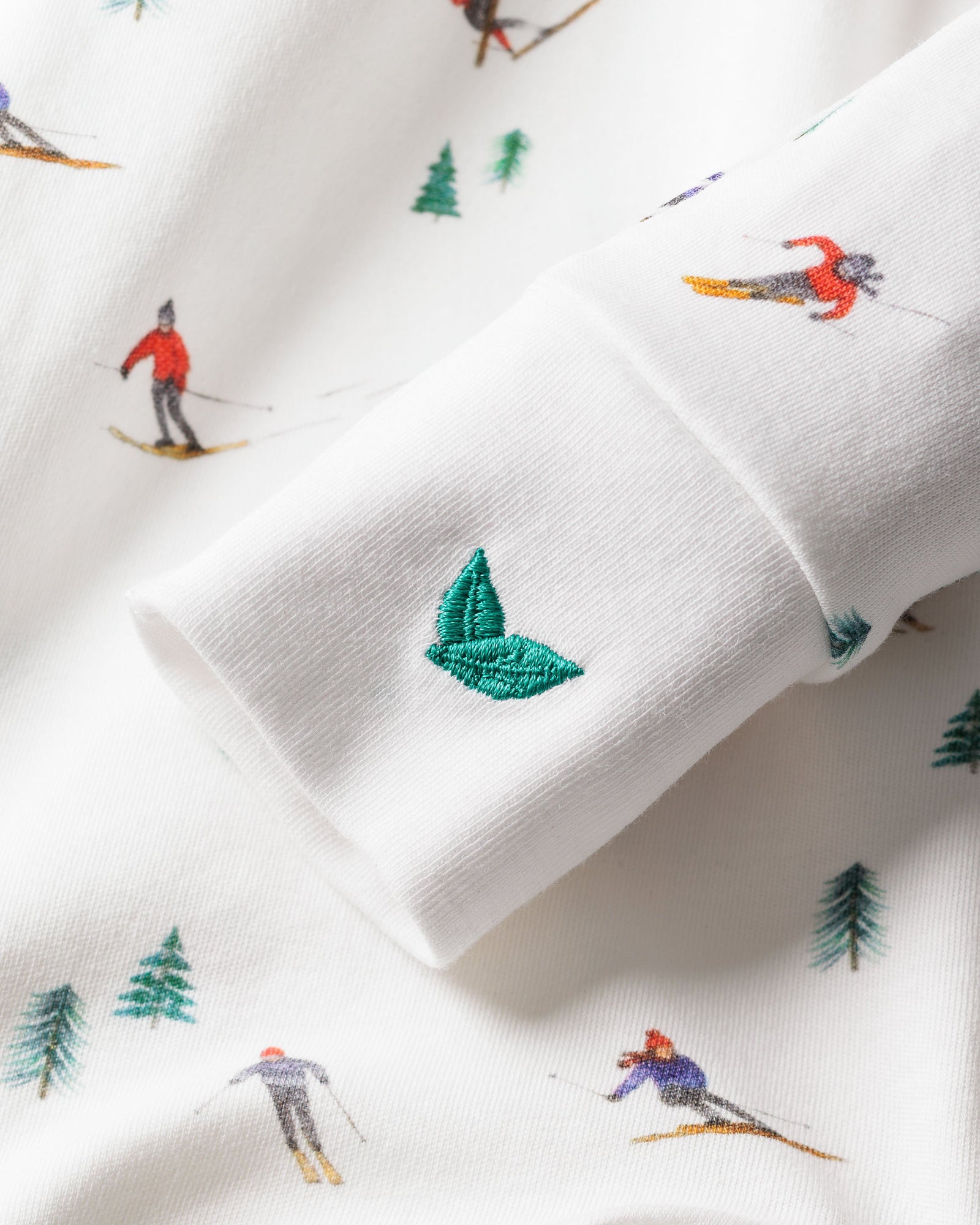 Close-up of Petite Plumes Babys Pima Romper in Apres Ski, showcasing a small embroidered green leaf on soft white Pima cotton fabric. The pattern includes skiers in vibrant red and blue attire with green pine trees. Leaf emblem is the focus.