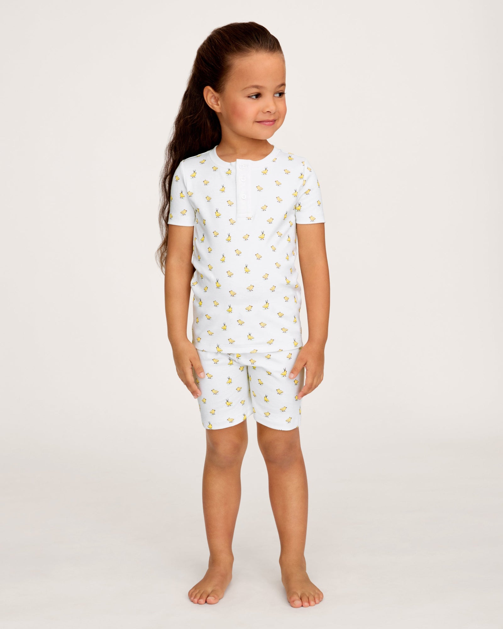 A young child with long brown hair smiles while wearing the Kid's Pima Snug Fit Short Set in Tres Chick, standing against a plain light background.