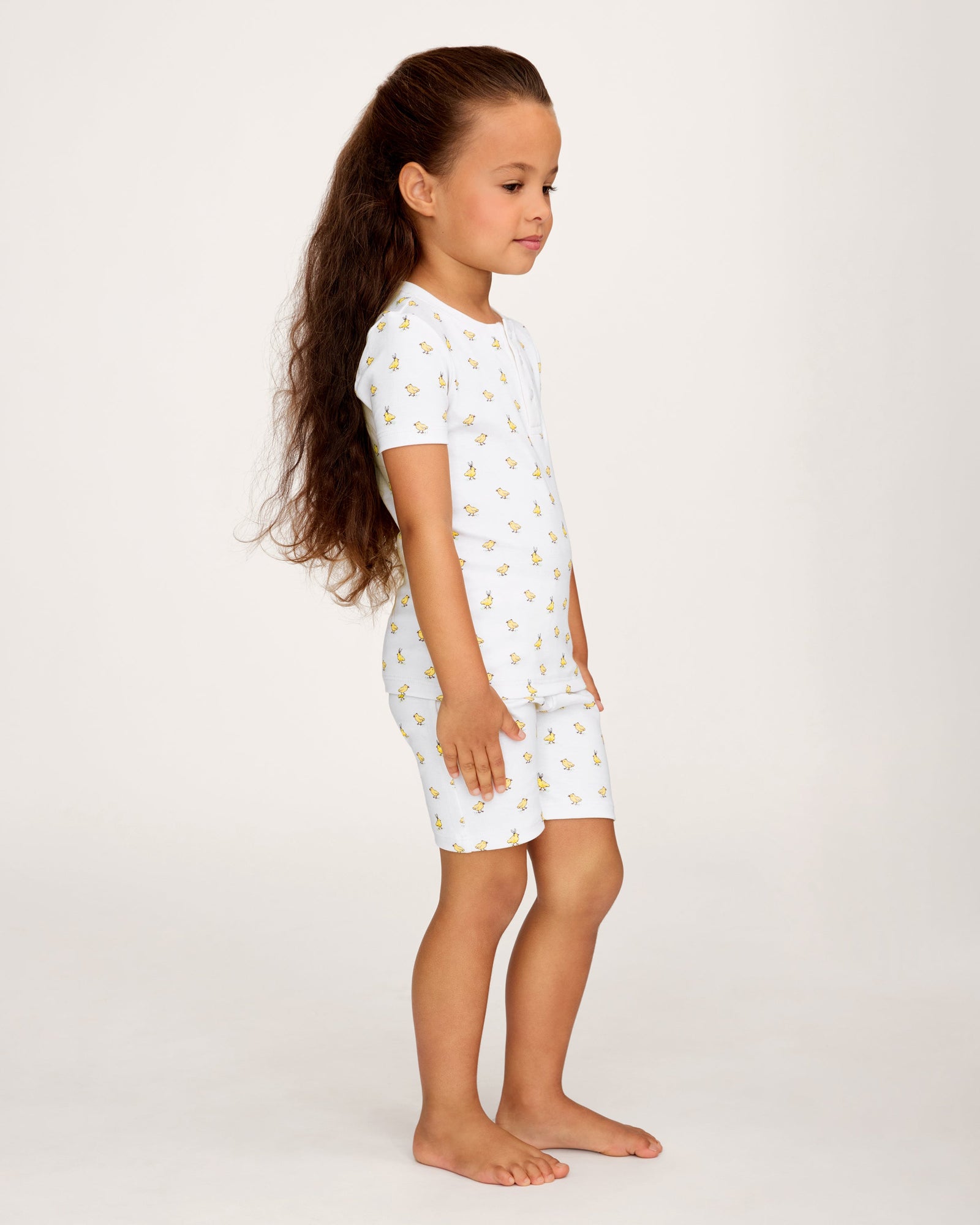 A young child with long brown hair smiles while wearing the Kid's Pima Snug Fit Short Set in Tres Chick, standing against a plain light background.