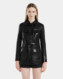 A woman with straight, long blonde hair models the Rudsak DANICA - 8125025 Black leather jacket, featuring four front pockets and buttons, paired with black shorts against a plain white background.