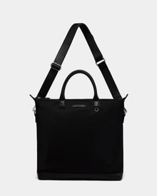 The DEXTER - 9324513 Black by Rudsak is a versatile unisex canvas tote with short handles, an adjustable shoulder strap, a rectangular shape, and a small metal nameplate on the front, shown against a plain white background.