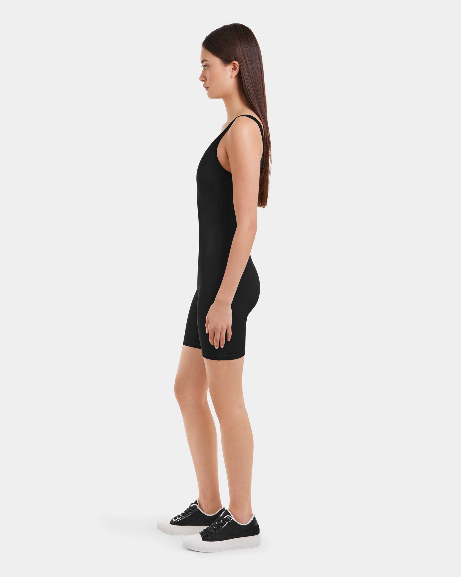 A woman stands in profile against a white background, wearing the Rudsak KAILEY - 8523012 Black Pattern fitted sleeveless romper, paired with black sneakers with white soles.