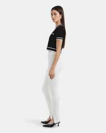A woman stands in profile against a white backdrop, wearing a black short-sleeve top with white trim, Rudsaks MATHILDA - 8524515 Iceland front ankle slit leggings in white, and black heeled shoes.